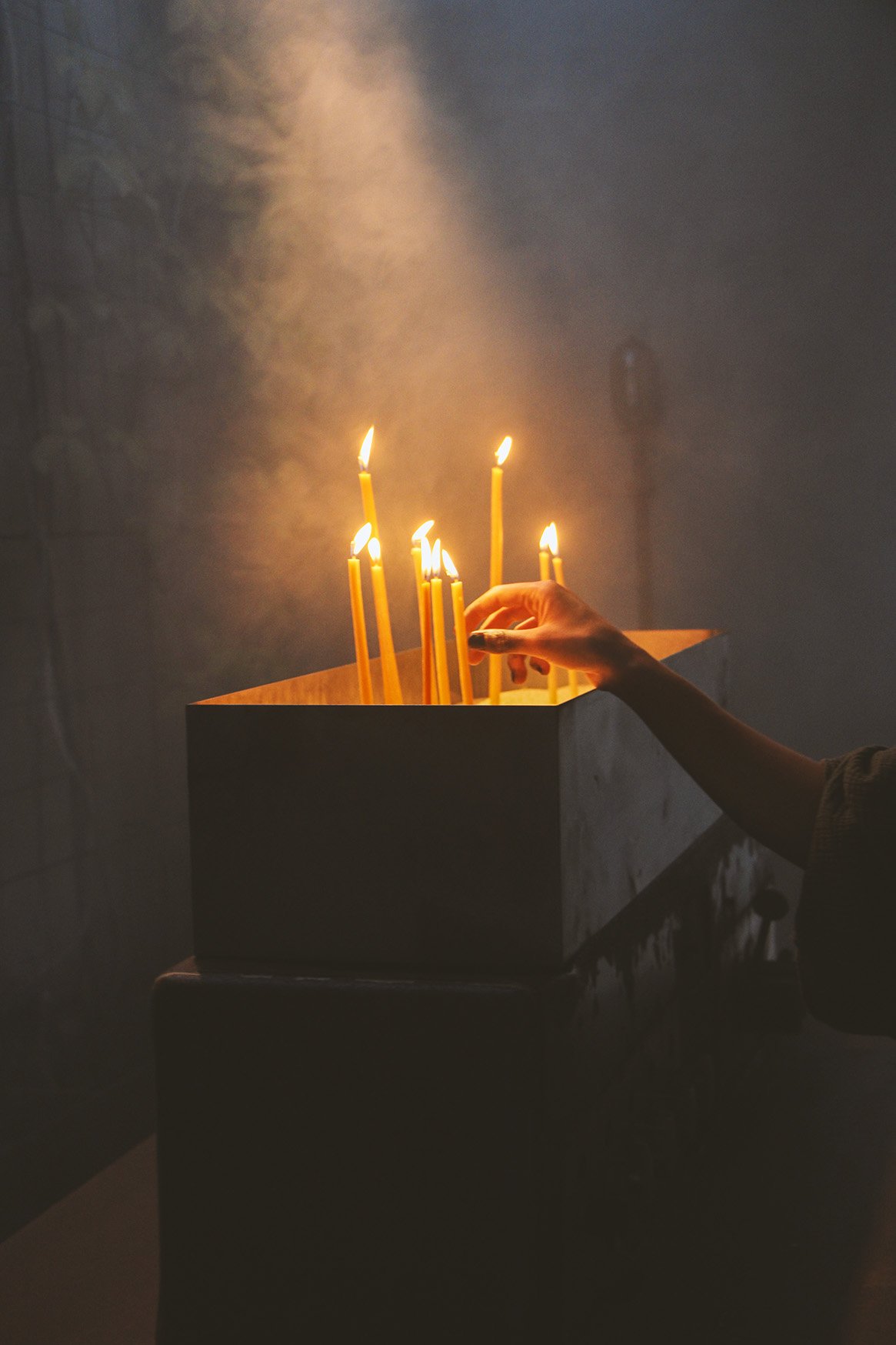 A person's hand lights tall candles in a dimly lit area with visible smoke.