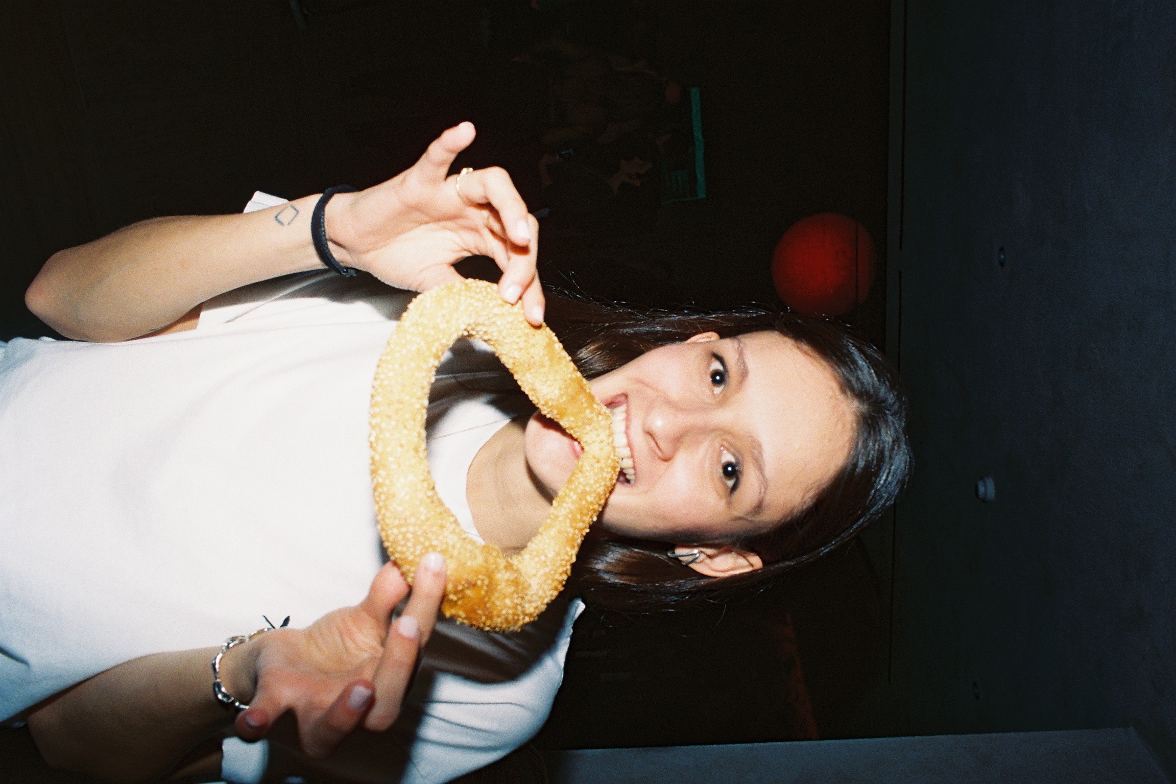 A smiling person in a dimly-lit room bites into a large, sesame-covered bagel. They wear a white shirt, creating a casual and joyful atmosphere.