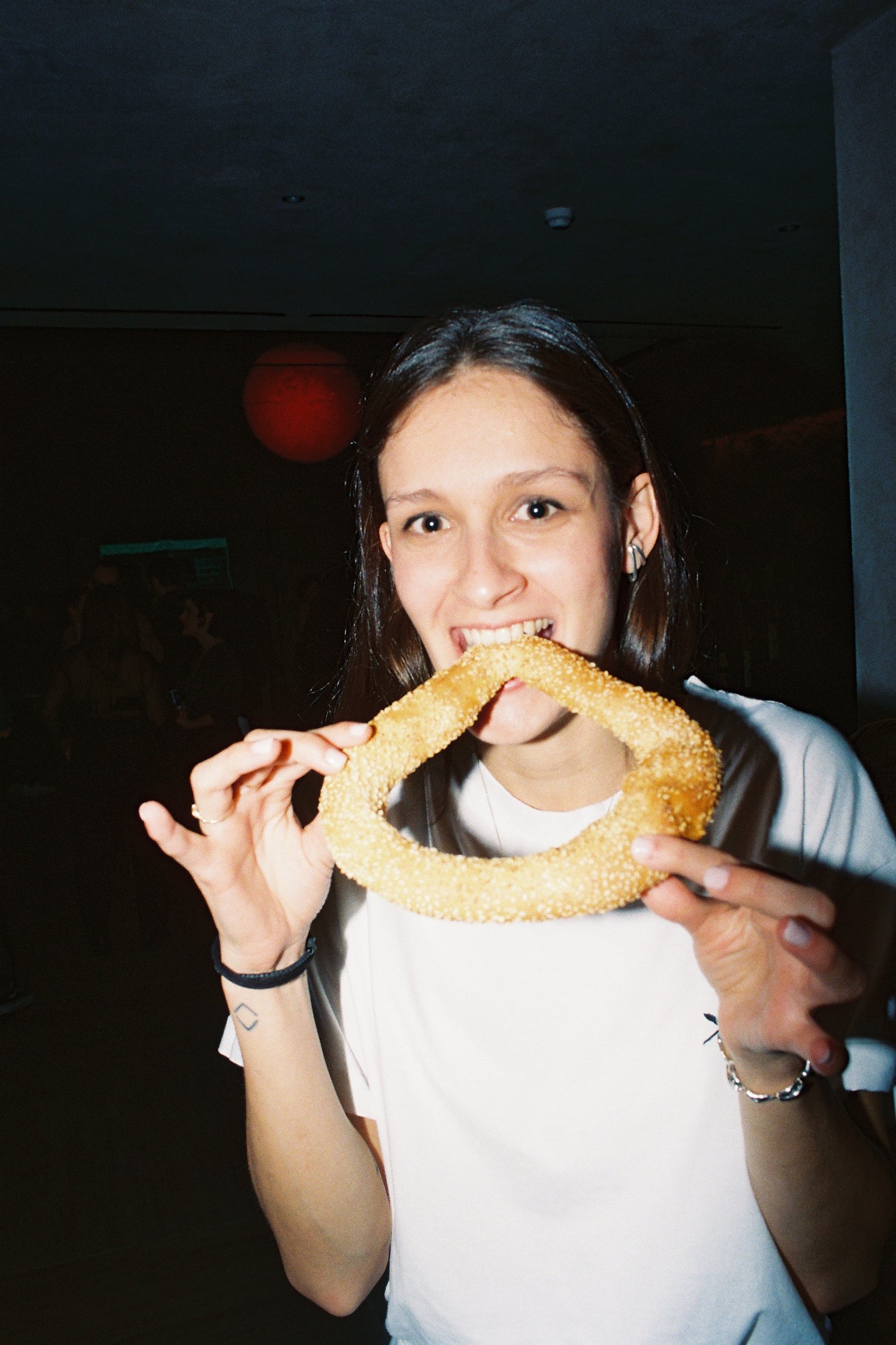 A smiling person in a dimly-lit room bites into a large, sesame-covered bagel. They wear a white shirt, creating a casual and joyful atmosphere.