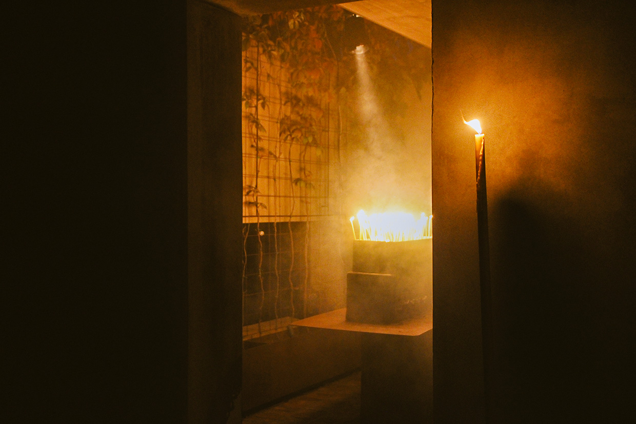 Lit candles in a dimly lit room cast a warm glow, creating a serene and ritualistic atmosphere amidst the haze.