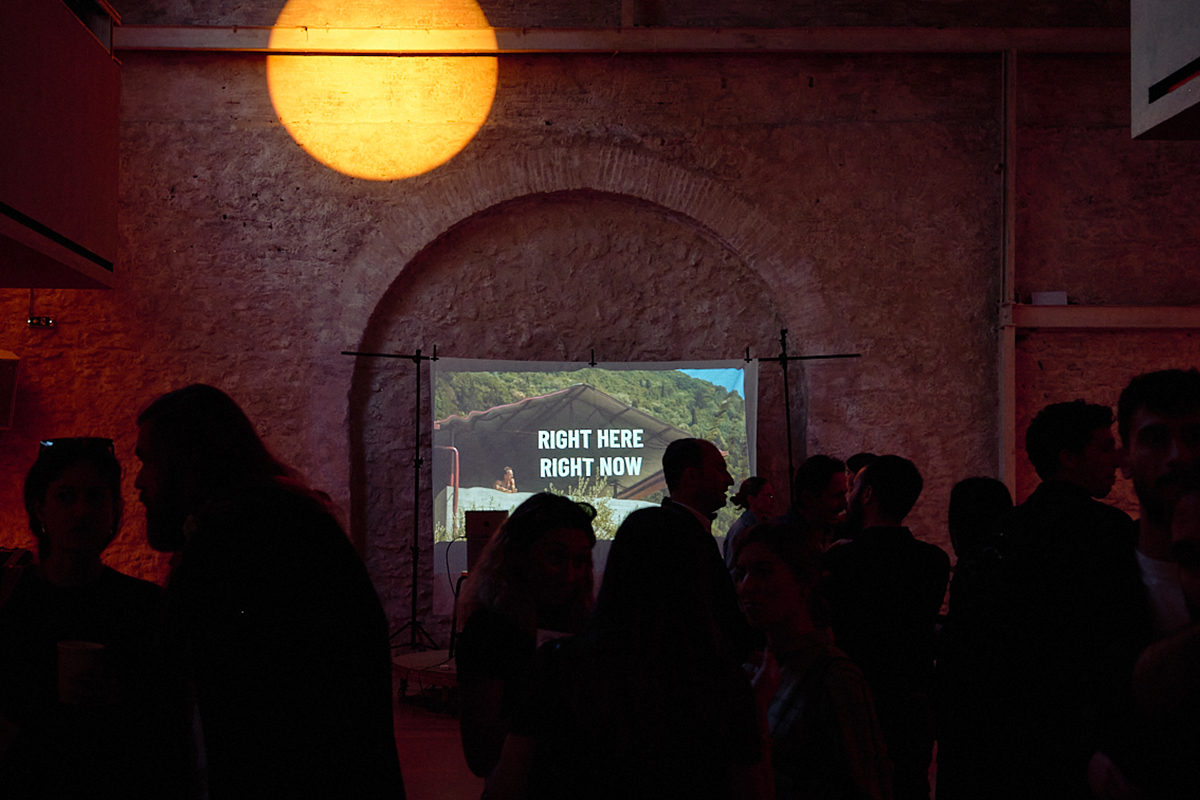 A crowd gathers in a dimly lit room with a rustic stone wall. A projector displays “RIGHT HERE RIGHT NOW,” casting a warm, inviting glow.